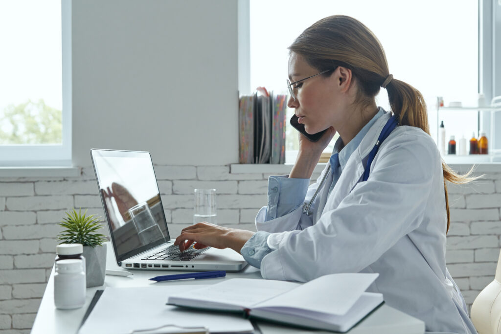 Confident female doctor talking on the phone while using her laptop to coordinate an outsourcing implementation plan